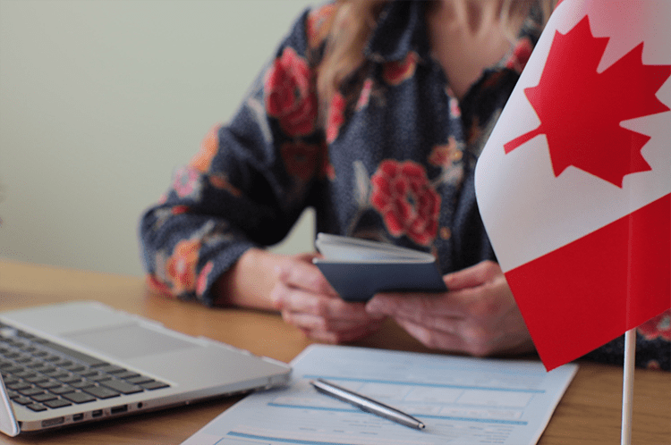 Canadian flag on the right hand side of the image, with a person sitting at a desk, holding a passport, with a laptop and some forms on the desk in front of her