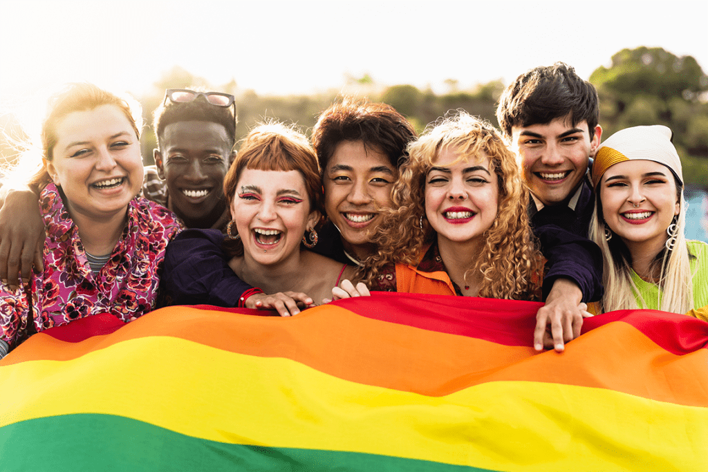 groupd of multi-racial queer young adults smiling together at the camera, holding a Pride flag in front of them