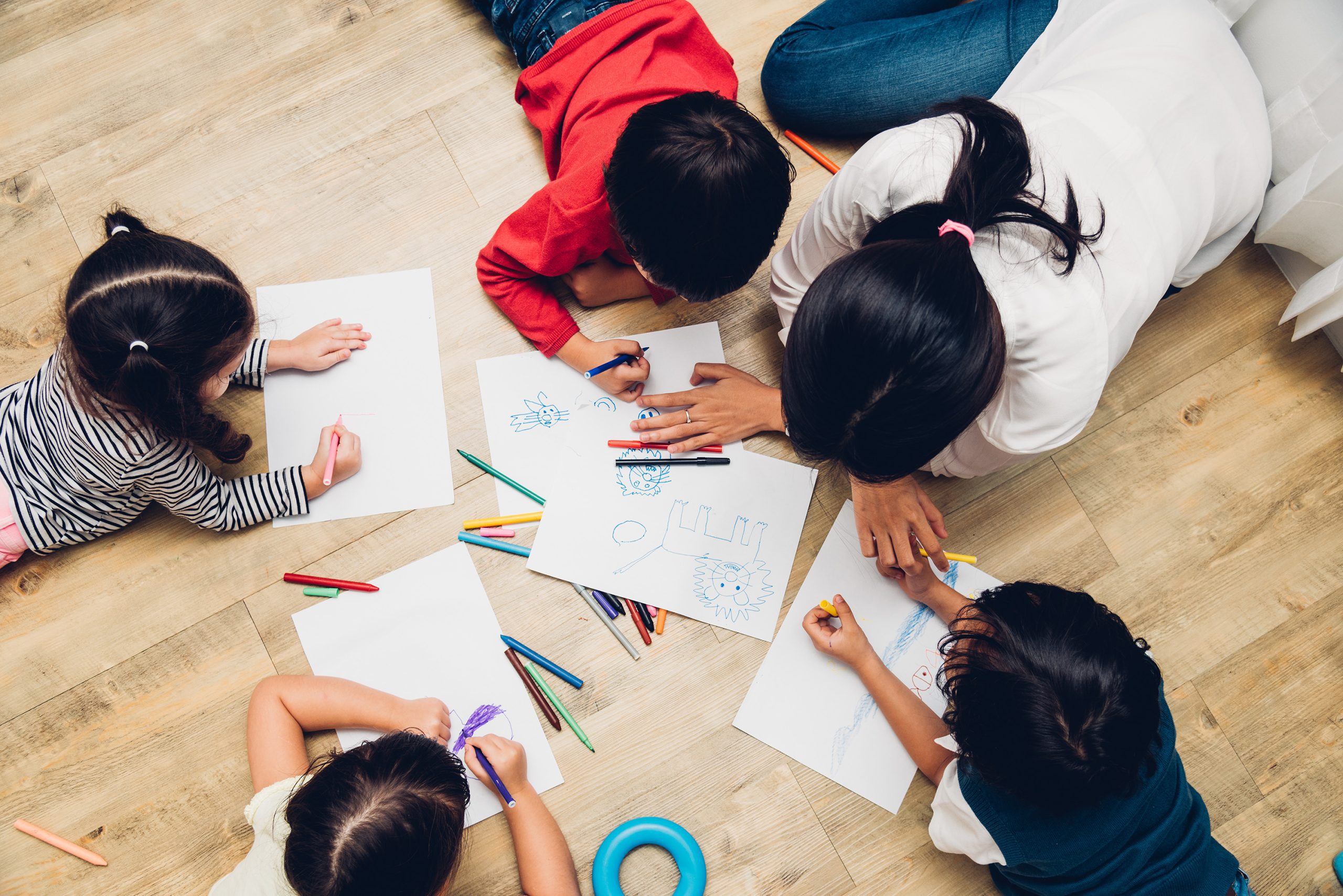 woman and 4 children with balck hair drawing pictures on white paper on a light brown wood floor, shot from above
