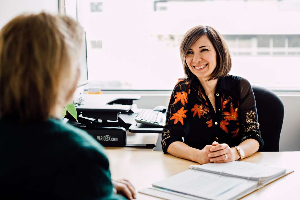professional woman sitting at a desk, smiling, speaking with an elderly woman on the other side of the desk, whose back is to the camera